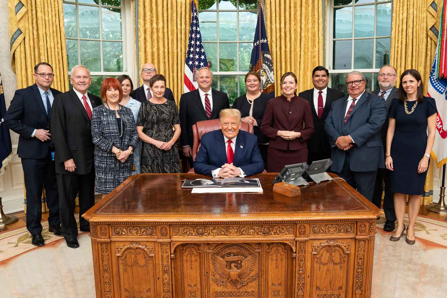 Kuvassa presidentti Donald Trump keskellä istumassa työpöytänsä takana Valkoisessa talossa ja taustalla seisomassa luovutusta saattanut delegaatio.