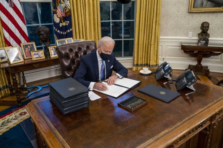 Joe Biden istuu työpöytänsä takana Oval Officessa ja allekirjoittaa päätöksiä.