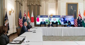 Joe Biden istuu pöydän takana esikuntansa istuessa etäänpänä. Ruuduilla kokoukseen osallistuvat Japanin, Intian ja Australian johtajat maidensa lippujen ympäröiminä.
