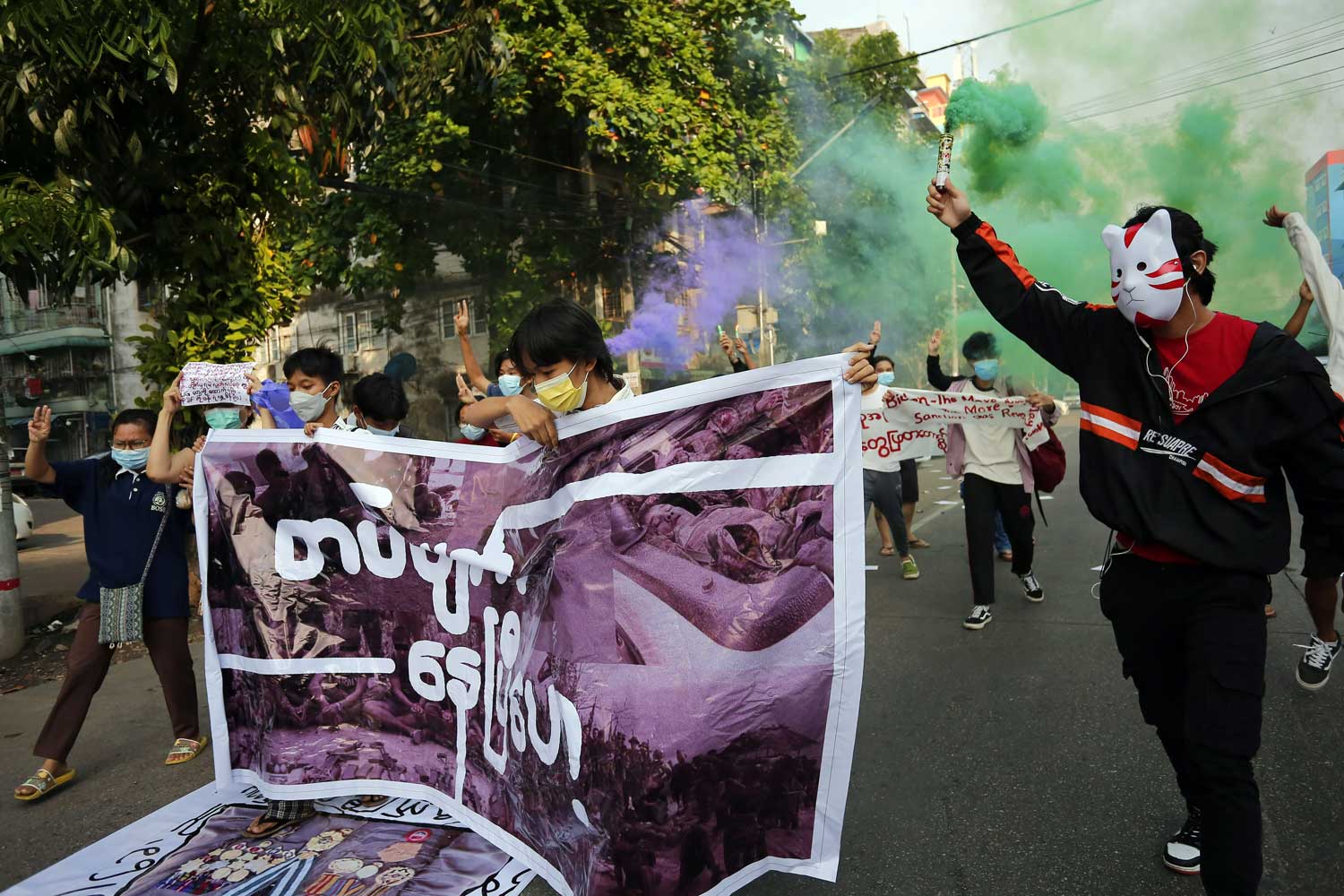 Mielenosoittajia kadulla banderollin, värisavujen ja naamareiden kanssa.