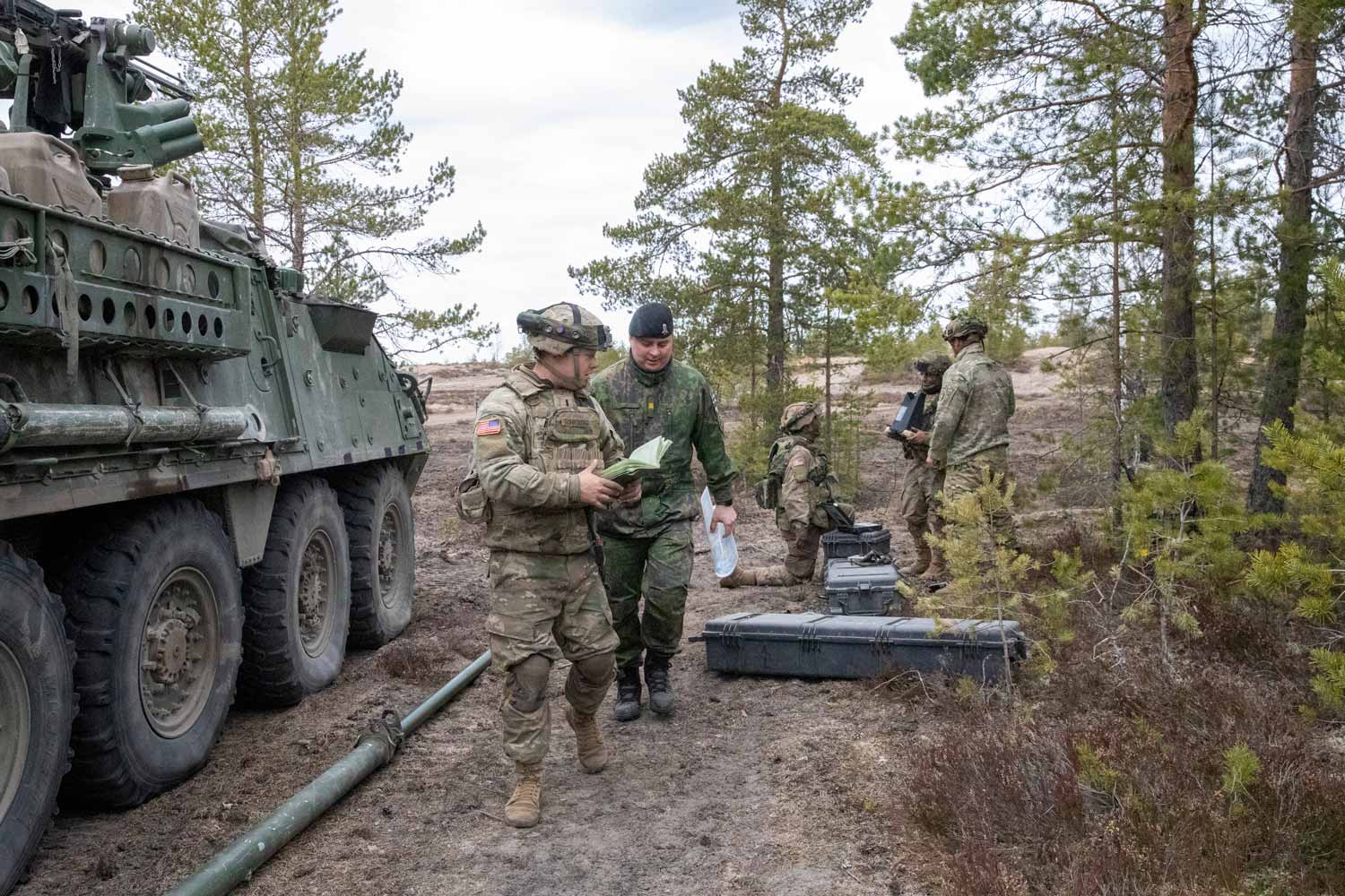 Vasemmalla panssaroitu ajoneuvo ja oikealla yhdysvaltalainen ja suomalainen aliupseeri kävelevät paperit kädessä.