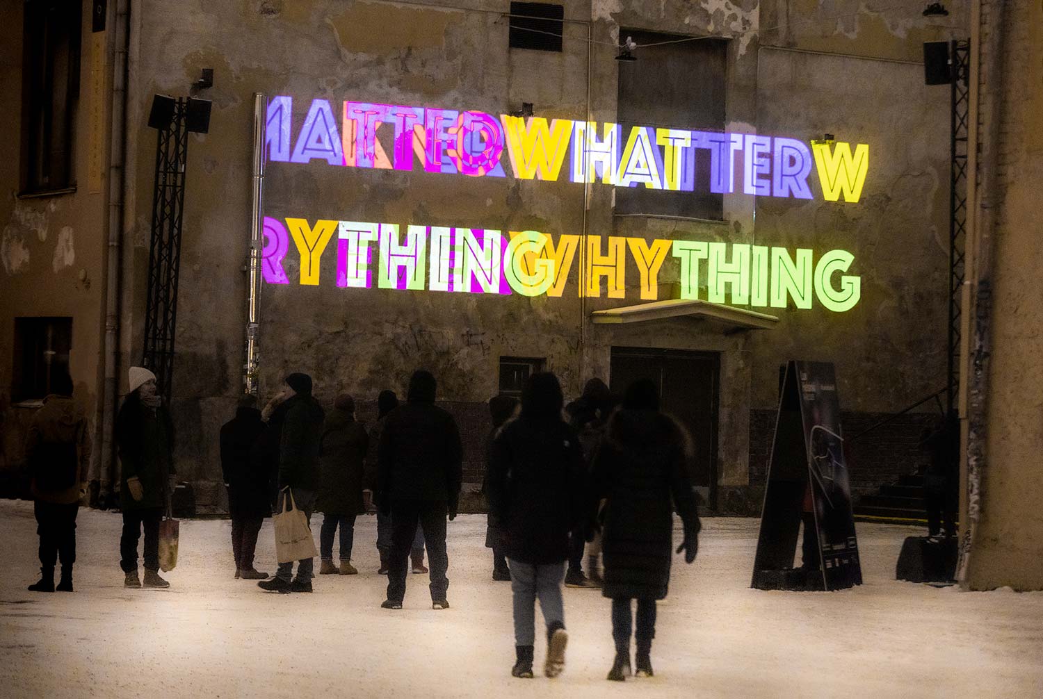 Ihmiset katsovat valoinstallaatiota, jossa seinälle heijastuu eri värisiä sanoa, kuten what, matter, anything, everything.