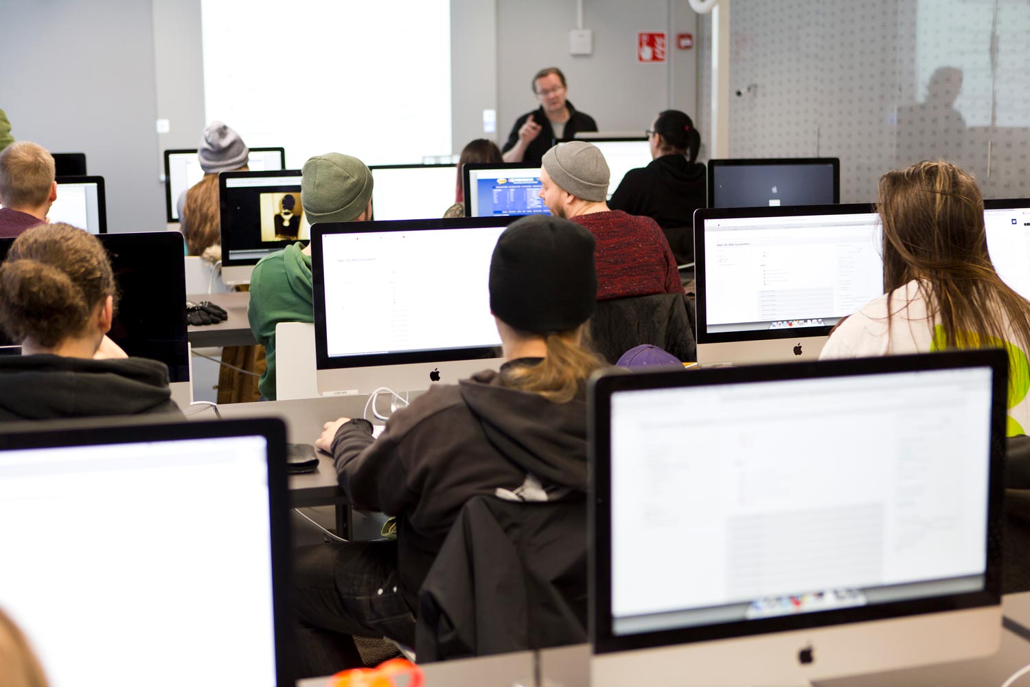 Opettaja puhuu luokan edessä. Pöydään ääressä istuvilla oppilailla on edesään tietokoneen ruudut, joista valtaosa loistaa valkoisena.