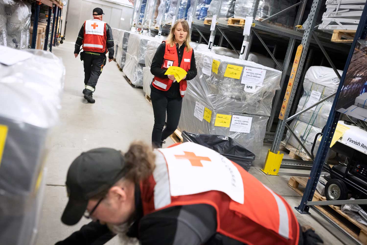 Kolme Punaisen Ristin liiveissä kulkevaa ihmistä töissä varastohallissa.