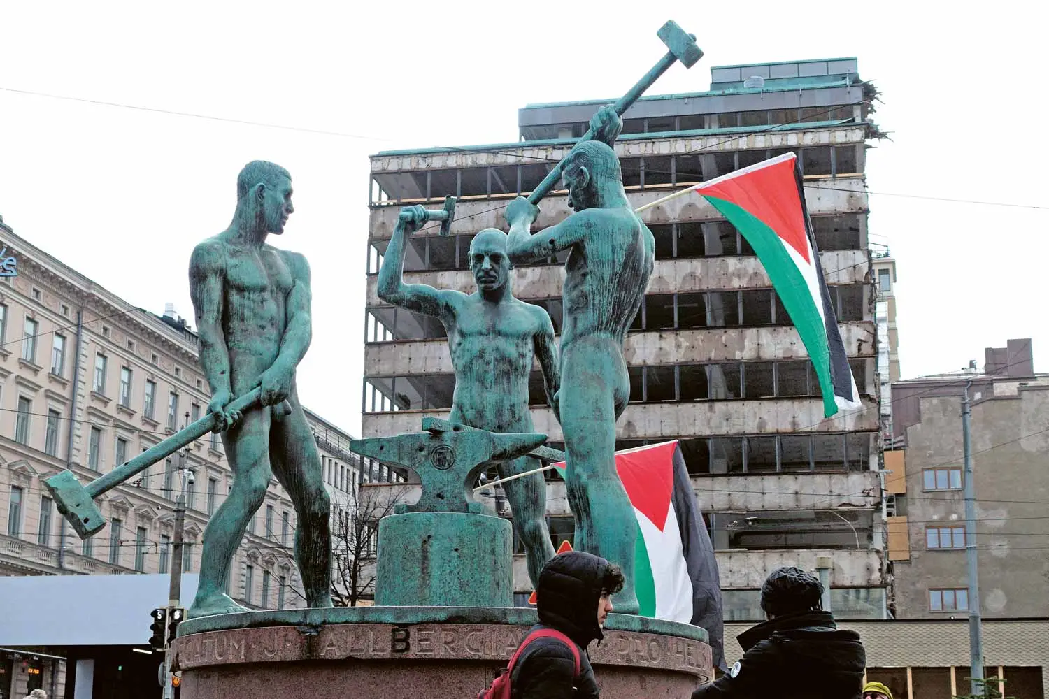 Kuvassa Kolmen sepän patsaaseen on ripustettu kaksi Palestiinan lippua. Taustalla käynnissä rakennuksen purkutyöt.