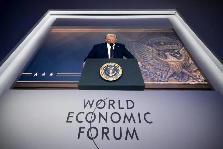 Presidentti Donald Trump puhuu puhujakorokkeen takana suoralla videoyhteydellä. Video on heijastettu seinälle ja sen alla lukee World Economic Forum.