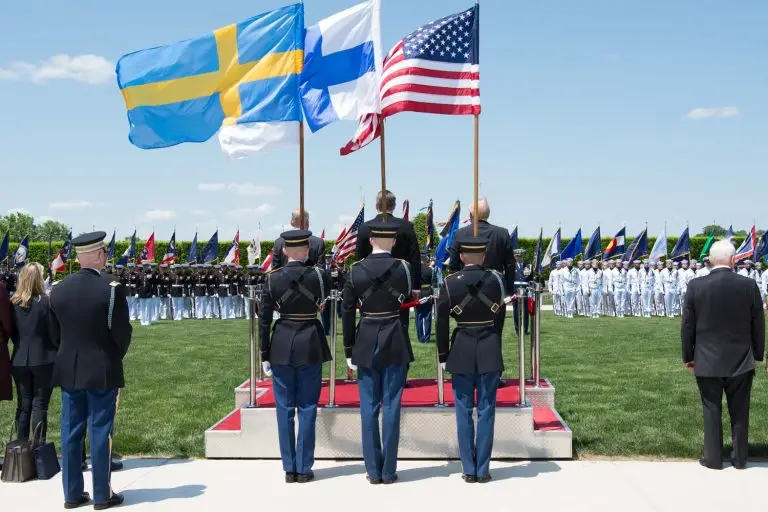 Puolustusministerit Peter Hultqvist, Jussi Niinistö ja Jim Mattis seisovat paraatikentällä selin kameraan Ruotsin, Suomen ja Yhdysvaltojen lippujen heiluessa kesäisessä säässä.