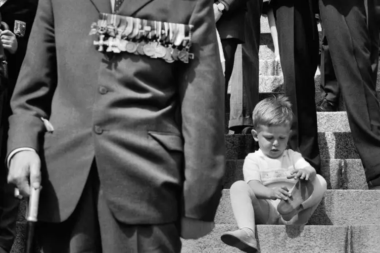 Mustavalkovalokuva on rajattu niin, että portailla istuvan kenkiä tutkailevan pojan etupuolella seisoo mies puvussa kävelykepin kanssa ja hänen pitkä kunniamerkkirivinsä asettuu kuvan etureunaan. Poikaa ympäröivien miesten kasvoja ei näy.