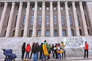 Kourallinen ihmisiä osoittaa mieltään Eduskuntatalon portaiden edessä. Yhdellä on hartioilla Katalonian lippu. Tuuli on taittanut kulman banderollista, josta on näkyvillä teksti Vapaus Katalonian poliittiset.