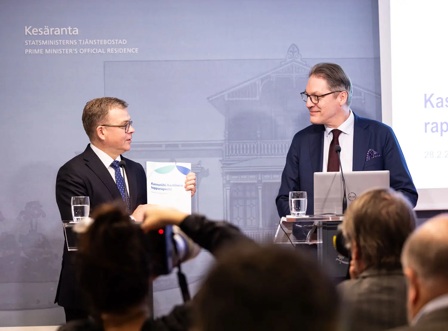 Pääministeri Petteri Orpo pitää käsissään Kasvuriihi-raporttia ja katsoo vieressä seisovaa Risto Murtoa raportin tiedotustilaisuudessa median edessä.