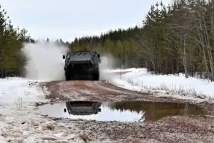 Panssaroidun miehistönkuljetusvaunun pyörät lennättävät vettä lumenreunustamalla lätäkköisellä soratiellä metsän keskellä.