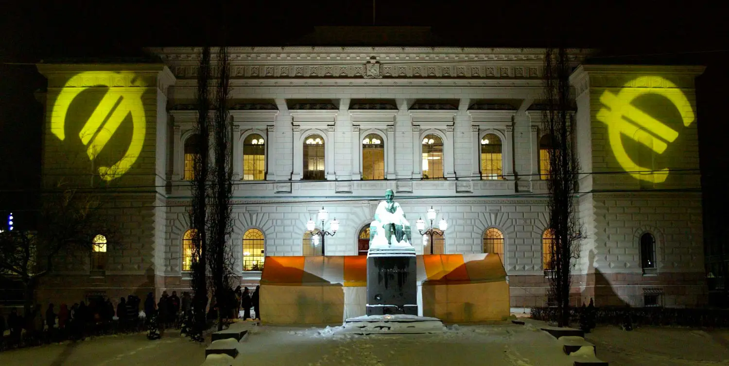 Öinen Suomen Pankin päärakennus on valaistu kirkkaasti keskeltä ja valaisematta jätetyihin rakennuksen kulmiin on heijastettu keltainen euro-symboli. Talvisen maiseman keskellä kulkee jono rakennuksen pääovelle.