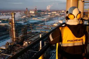 Jalostamon tornissa seisova henkilö katsoo talvista jalostamon maisemaa turvakypärä päässä ja Rosneft-tekstillä varustettu takki päällä.